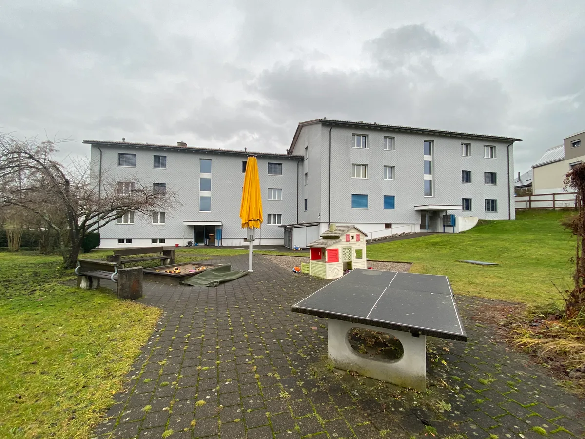 Zwei zusammengebaute Mehrfamilienhäuser. Im Vordergrund ein Spielplatz mit Tischtennistisch, Sandkasten und einem kleinen Schloss aus Kunststoff.
